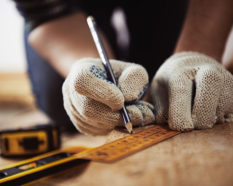Close-up of craftsman hands in protective gloves measuring wooden plank with ruler and pencil. Woodwork and renovation concept. Close-up of craftsman hands in protective gloves measuring wooden plank with ruler and pencil. Woodwork and renovation concept.
